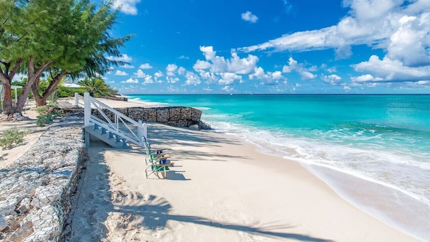 Una playa con agua turquesa cristalina, una escalera blanca que conduce a una terraza de madera y algunas sillas de playa.