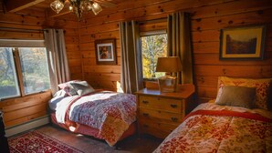 Een gezellig slaapkamer in een houten chalet met twee bedden, een kaptafel, een nachtkastje, een lamp en een raam met uitzicht op herfstbomen.