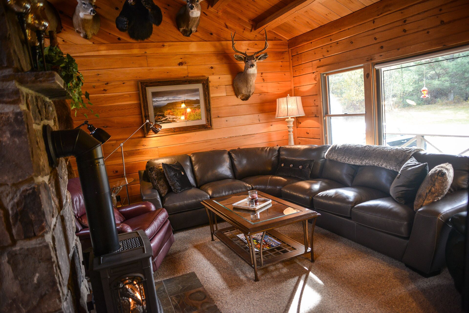 A cozy cabin interior with a leather sofa, a glass coffee table, a fireplace, and a deer head mounted on the wall.