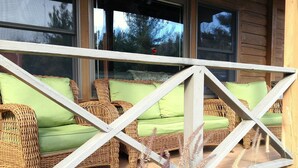 Een rieten bank met groene kussens op een houten terras, met een houten leuning op de voorgrond.