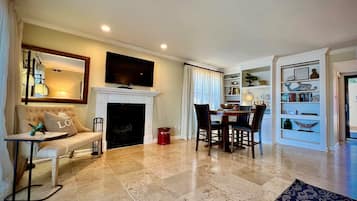 A living room with a fireplace, a television mounted above it, a dining area with a table and chairs, and a bookshelf with decorative items.