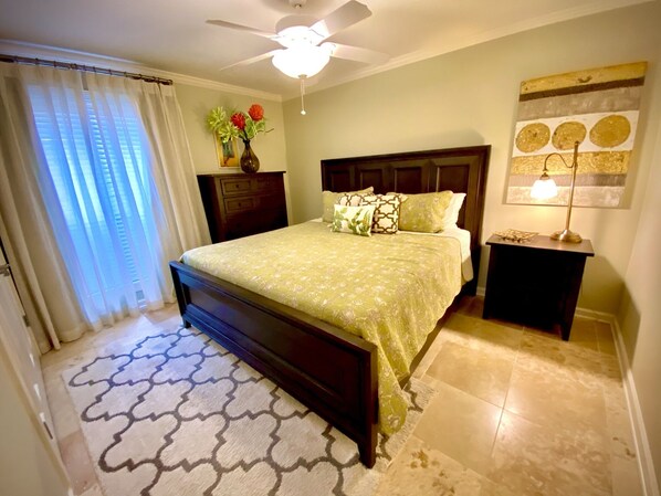 A bedroom with a bed, a nightstand, a lamp, a ceiling fan, a window with curtains, and a vase with flowers.