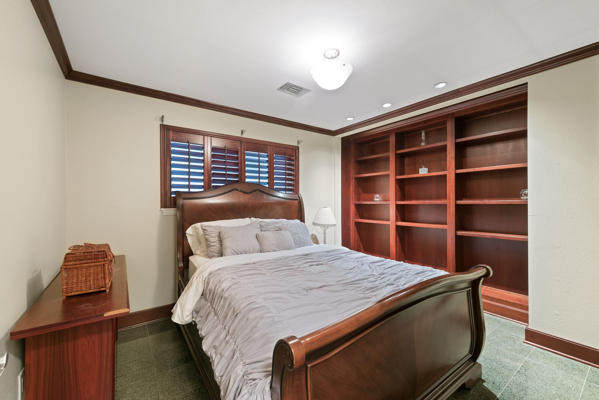 Een slaapkamer met een houten bed, een nachtkastje, een rieten mand, een raam met zonwering en ingebouwde houten planken.