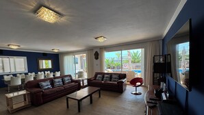 A spacious living room with two sofas, a coffee table, and a TV. There is a dining area with white chairs and a table. Large windows offer a view of a pool and outdoor area. - Detached Villa with Private Pool, close to all amenities and a blue flag beach (Santiago del Teide)