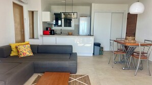 A modern living room with a grey sofa, a wooden coffee table, and a dining area with metal chairs and a wooden table. - sea ​​view Apartment Direct access private beach, swimming pools, sauna, hammam, tennis etc. (Théoule-Sur-Mer)