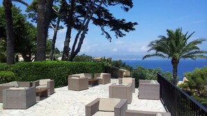 An outdoor seating area with wicker furniture and a stone floor, overlooking a scenic view of the sea. - sea ​​view Apartment Direct access private beach, swimming pools, sauna, hammam, tennis etc. (Théoule-Sur-Mer)