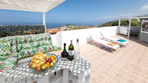 A terrace with a table set with fruits and bottles, lounge chairs, and a view of the sea. - Cozy studio with breathtaking sea and Teide views, quiet neighborhood. (LA GUANCHA)