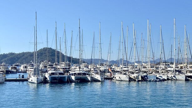 A marina filled with numerous sailboats docked in calm waters, surrounded by hills.