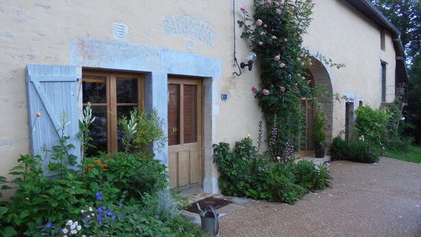 Un bâtiment doté de deux portes en bois, d’un petit jardin agrémenté de diverses plantes et fleurs, et d’un chemin de pierre menant à l’entrée.