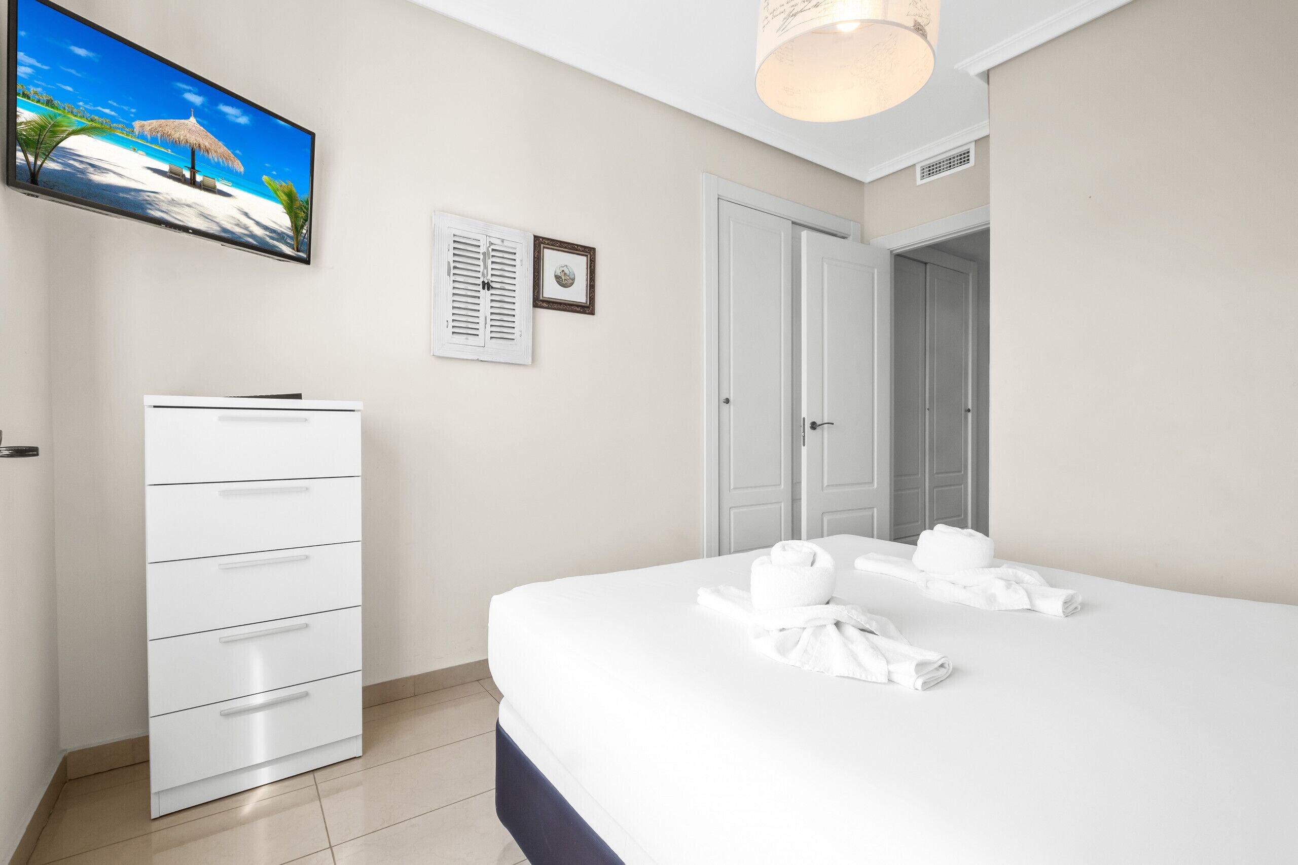 Une chambre d’hôtel avec un lit, une armoire, un téléviseur diffusant une scène de plage, une porte et un luminaire au plafond.
