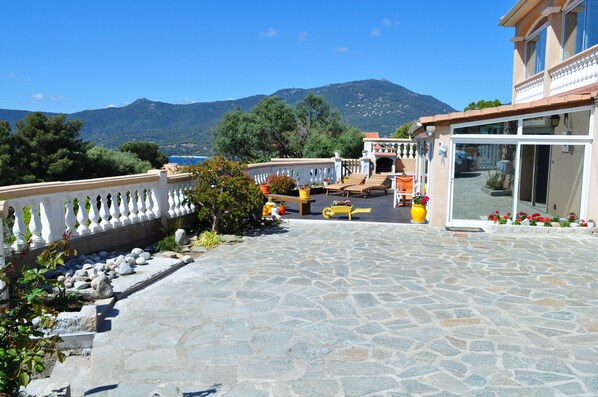 A stone-paved patio with outdoor furniture, a white balustrade, and a view of a mountain and sea.