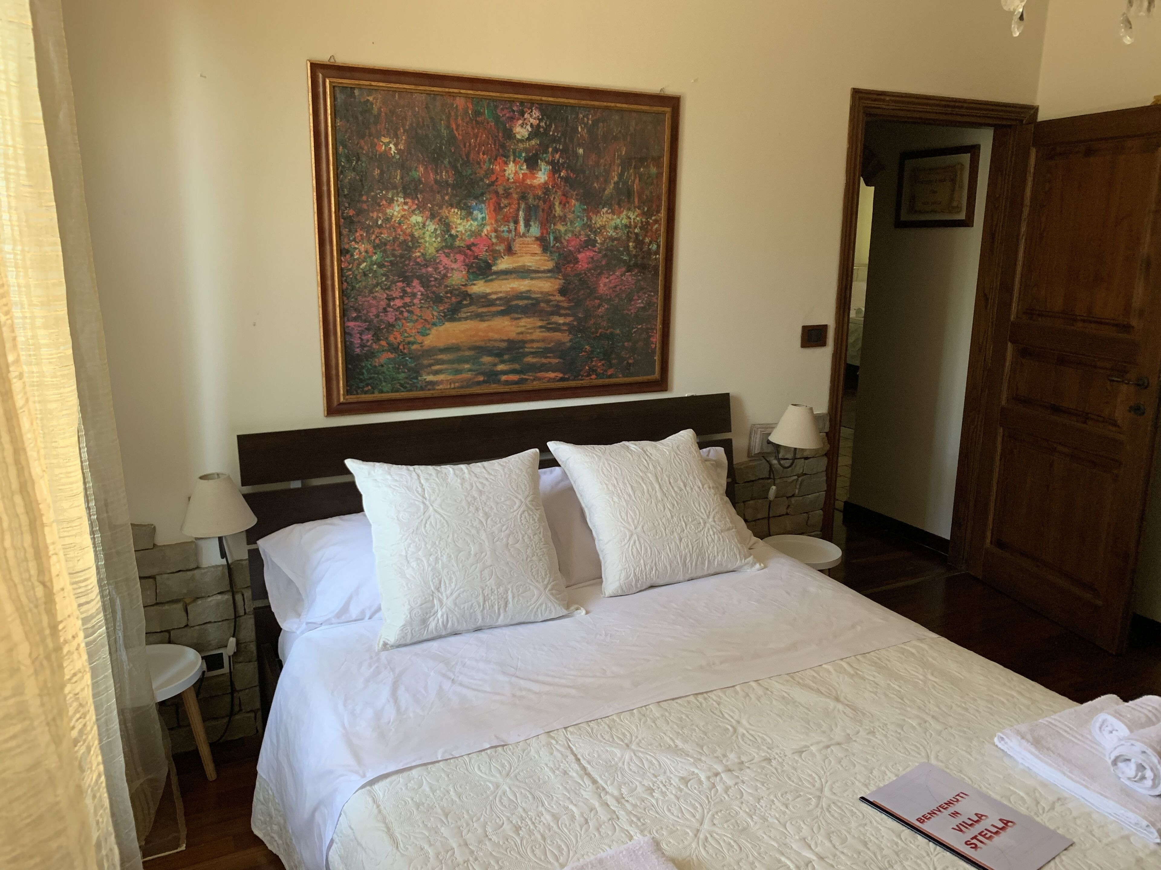 Habitación de hotel con una cama grande, ropa de cama blanca, un cuadro en la pared, una puerta de madera y una mesita con una lámpara.