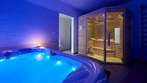 Un spa moderno con piso calefaccionado, una sauna con paredes de vidrio y una bañera de hidromasaje iluminada con luz azul.
