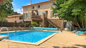 Una piscina con un muro de piedra, una zona de estar cubierta y una escalera que lleva a un balcón.