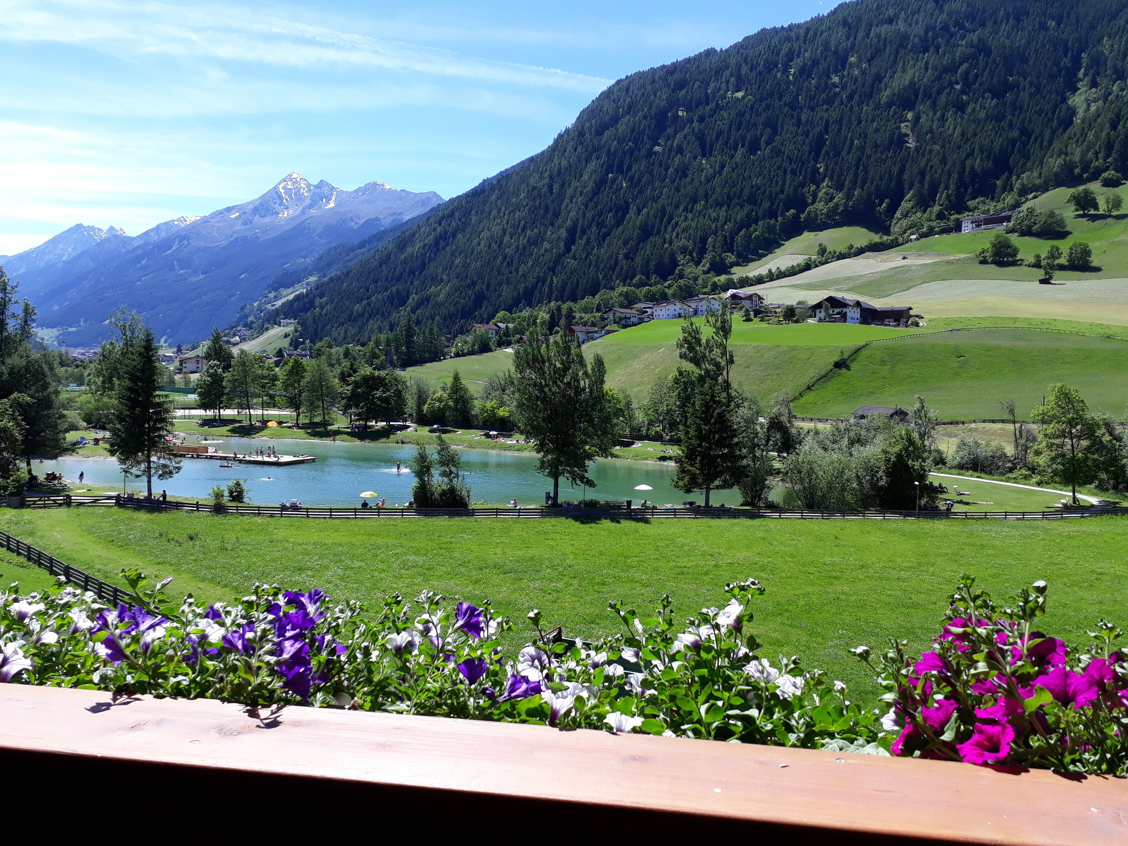 Una vista panorámica de un lago rodeado de montañas, con una terraza de madera en primer plano y un cielo despejado.