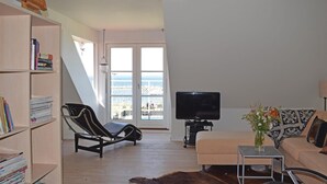 Ein Wohnzimmer mit Fernseher, einer Couch, einem Sessel, einem Bücherregal und einem großen Fenster mit Meerblick.