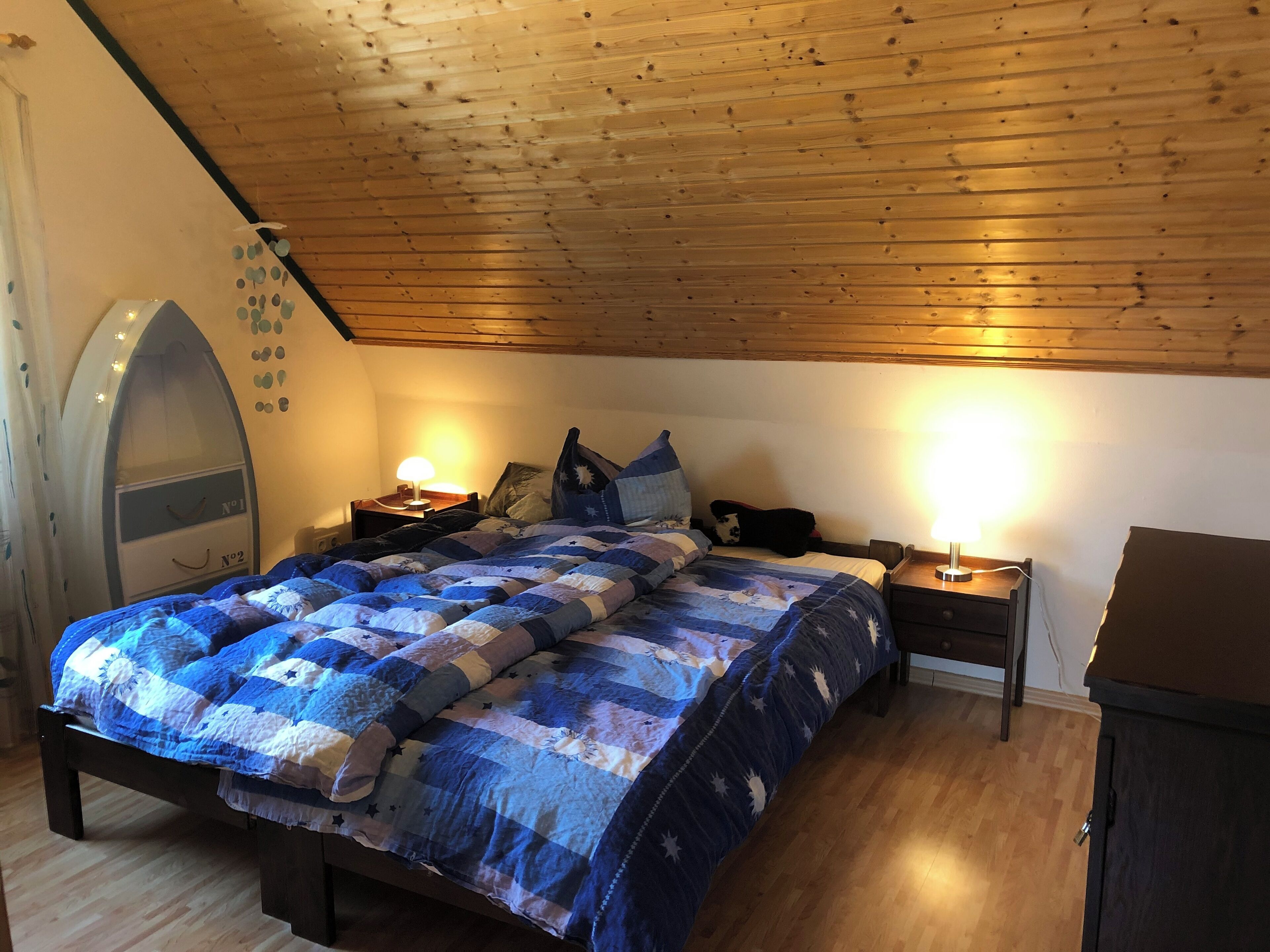 Ett sovrum med ett träpanelat tak, en säng med ett blått och vitt täcke, två sänglampor och en liten, välvd spegel på vänster sida.