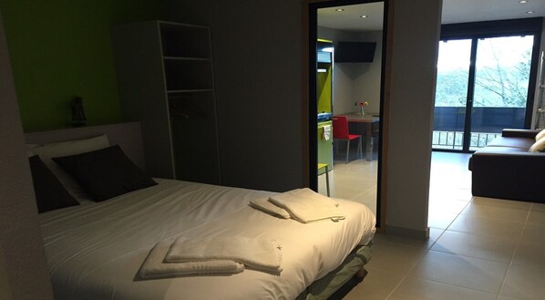 A modern hotel room with a large bed, a wardrobe, a balcony with a view, and a dining area visible through an open door.