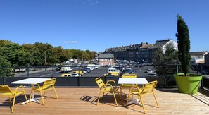A rooftop terrace with yellow chairs and tables, overlooking a busy street with cars and buildings.
