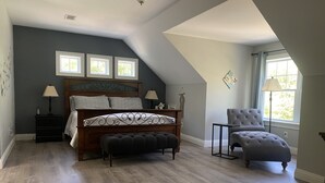 Een slaapkamer met een schuine plafond, een bed met hoofdeinde, een nachtkastje, een stoel en een raam met uitzicht.