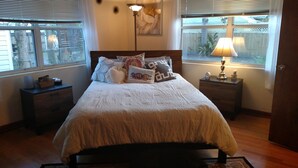 A bedroom with a large bed, multiple pillows, a nightstand with a lamp, and a window with blinds. - Comfy Safe Quiet Haven, Dog Friendly!  2 Miles to Downtown or to Vilano Beach (St. Augustine)