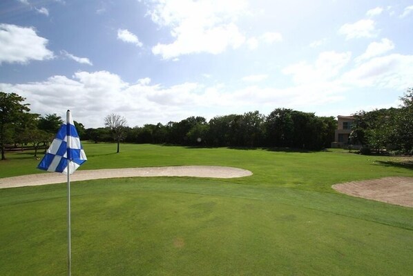 A golf course with a flag on a flagstick, a sand bunker, and a building in the background. - Quinta Del Sol, 3 Bedroom Beachfront Condo In Puerto Aventuras, Riviera Maya (puerto aventuras)