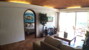 A living room with a sofa, a TV, a bookshelf, and a painting on the wall. - Quinta Del Sol, 3 Bedroom Beachfront Condo In Puerto Aventuras, Riviera Maya (puerto aventuras)