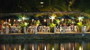 An outdoor dining area with tables and chairs, umbrellas, and a body of water in the foreground. - Quinta Del Sol, 3 Bedroom Beachfront Condo In Puerto Aventuras, Riviera Maya (puerto aventuras)