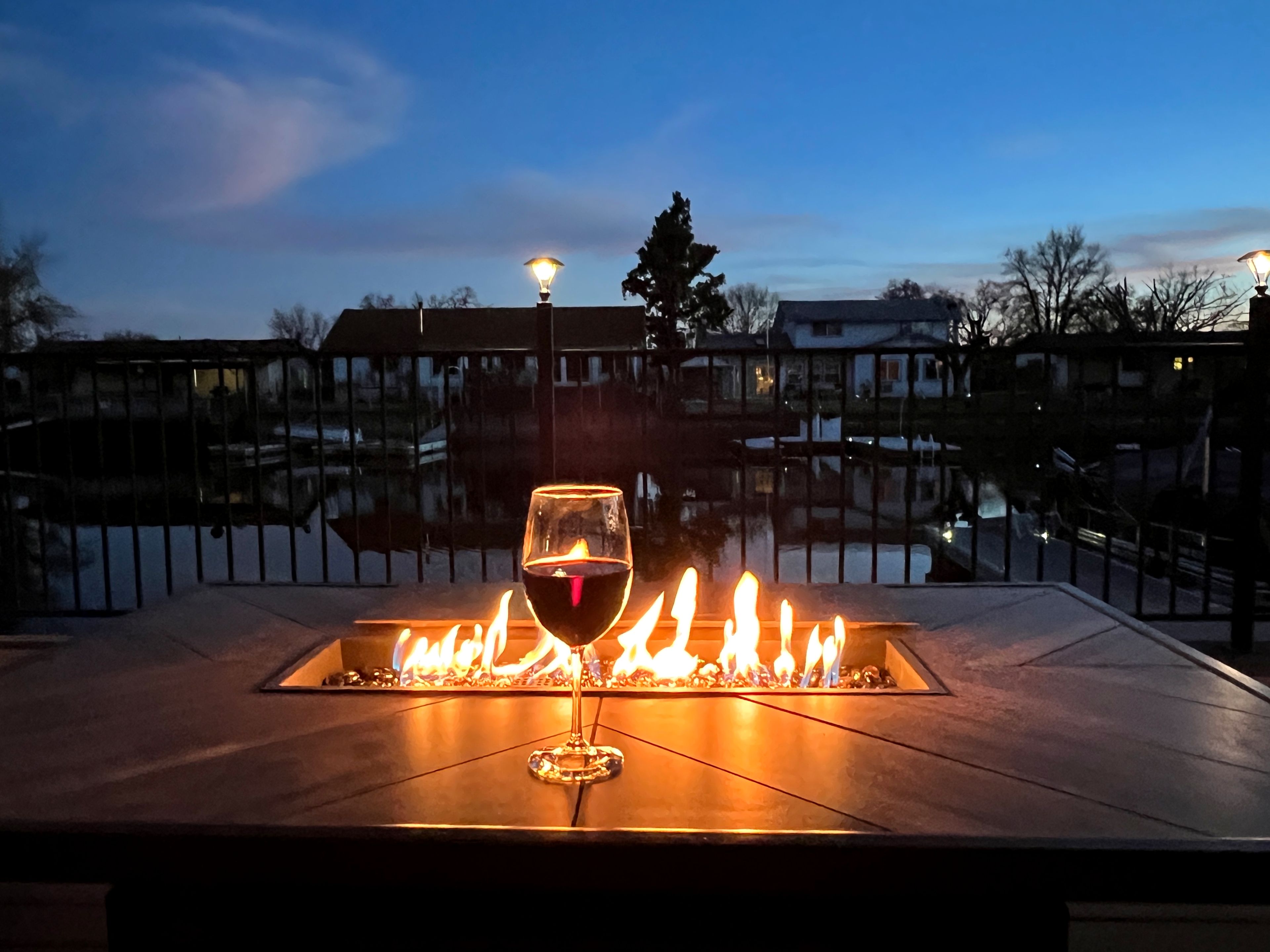 A glass of red wine on a fire pit with a view of a residential area at dusk.