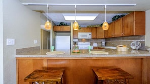 A kitchen with wooden cabinets, a central island with stools, hanging pendant lights, and a microwave.