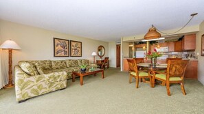 A living room with a sofa, coffee table, and dining table with chairs. There is a kitchen area visible in the background.