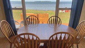 A wooden dining table with four chairs, set on a wooden floor, overlooking a grassy area with a body of water and a distant island.