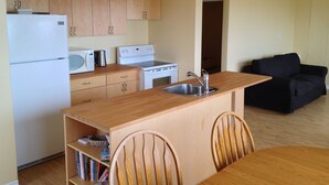 A compact kitchen with wooden cabinets, a white refrigerator, a microwave, and a stove. There is a dining table with chairs and a black sofa in the living area.