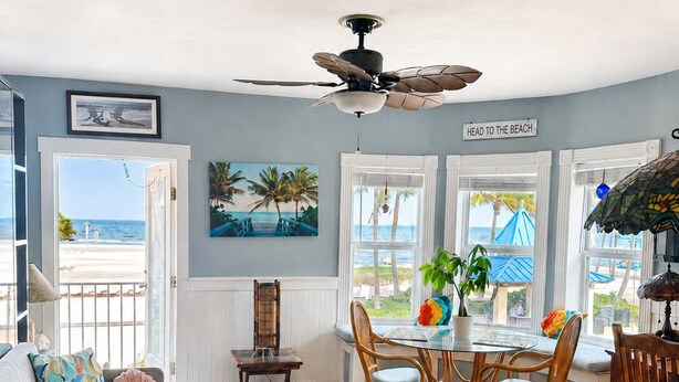 A room with a view of the beach, a ceiling fan, a dining table with chairs, and a sofa with cushions.