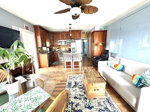 A modern kitchen with wooden cabinets, a dining area with a white table and chairs, a grey sofa with colorful cushions, and a ceiling fan.
