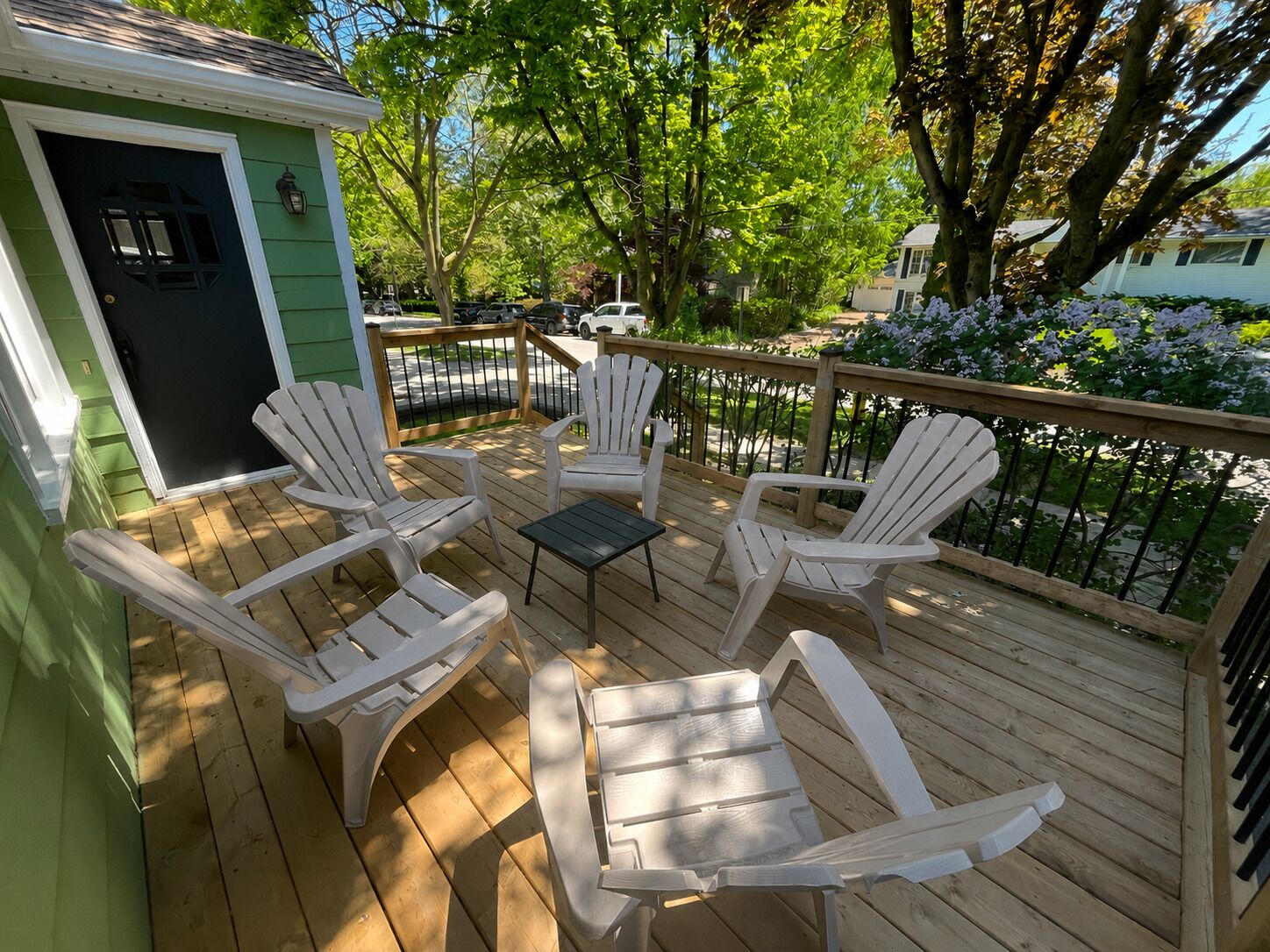 A wooden deck with white Adirondack chairs and a small table, surrounded by greenery and trees.