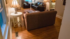 A living room with a brown sofa, two lamps, a small table, and a window with curtains. - The Lily Pad - quaint home in Old Town NOTL, steps from the lake! (Niagara-on-the-Lake)