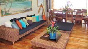 Ein Wohnzimmer mit einem gewebten Sofa, einem Couchtisch mit Pflanze und einem Essbereich mit Holztischen und Esstisch.