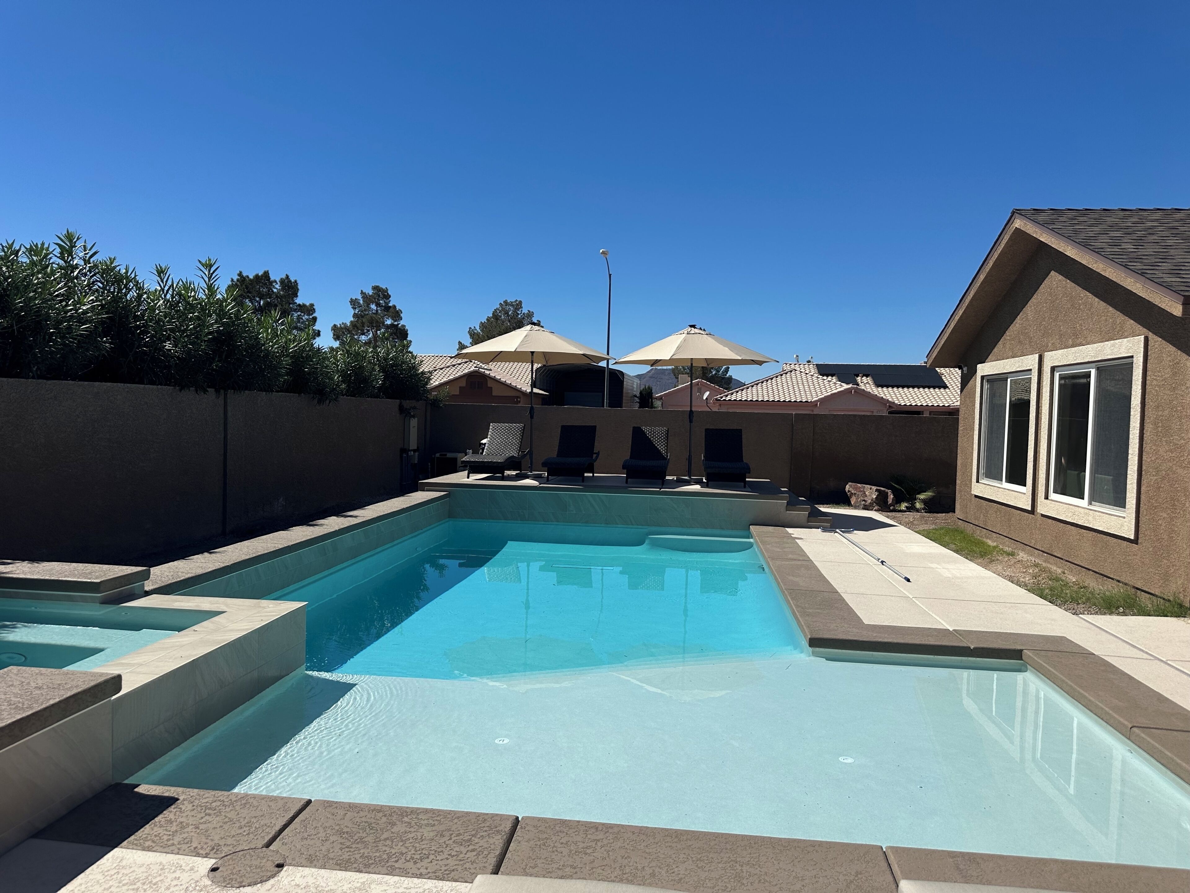 A swimming pool with a tiled deck, lounge chairs, umbrellas, and a house with a tiled roof in the background.