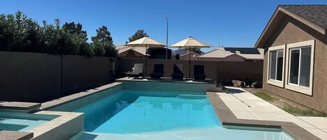 A swimming pool with a tiled deck, lounge chairs, umbrellas, and a house with a tiled roof in the background.