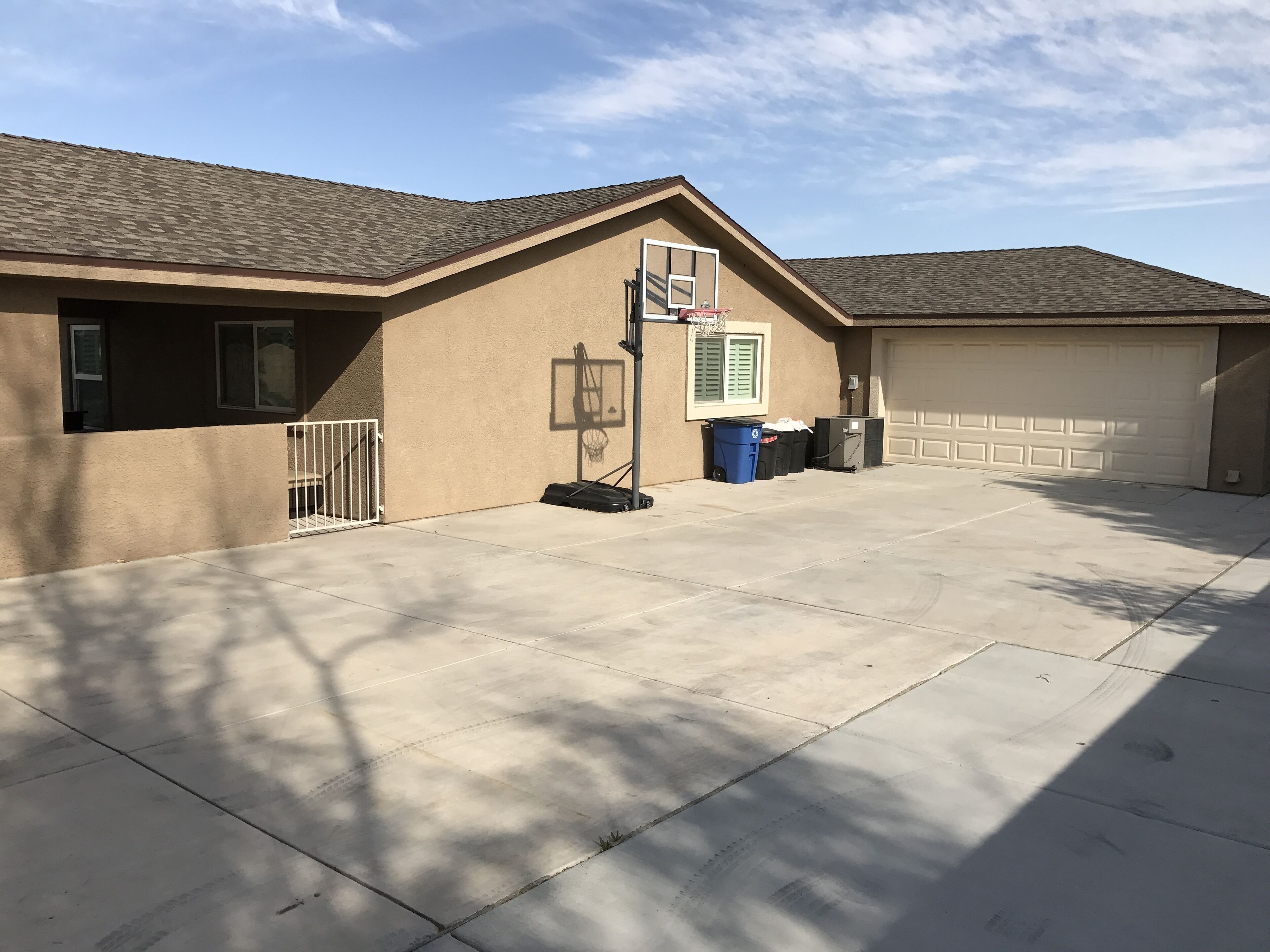 Un garage attenant à une maison, avec une allée en béton, un panier de basketball et une poubelle.