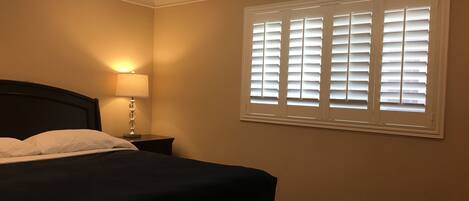 A bedroom with a bed, a nightstand with a lamp, a window with blinds, and a chandelier.