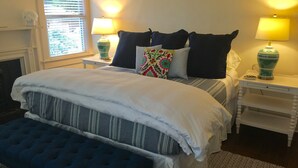 A neatly arranged bedroom with a large bed, two bedside tables with lamps, a window with blinds, and a fireplace.