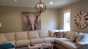 A living room with a beige sectional sofa, a wooden coffee table, a large wall clock, and a chandelier.