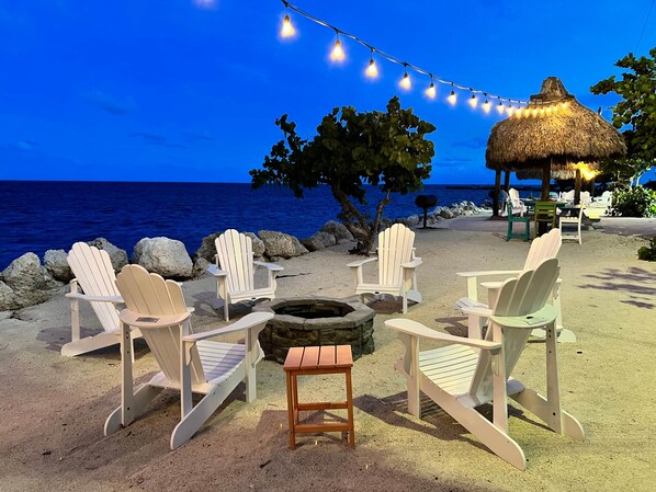 Un espacio de descanso frente a la playa con sillas Adirondack, una hoguera y luces colgantes.