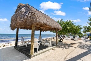 A beachside wooden deck with a thatched roof, white chairs, and a view of the ocean.