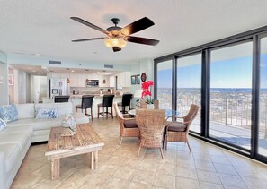 A spacious living area with a large window, a ceiling fan, and a kitchen in the background.