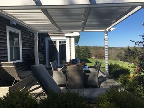 A covered patio with outdoor furniture, a grill, and a view of a grassy landscape.