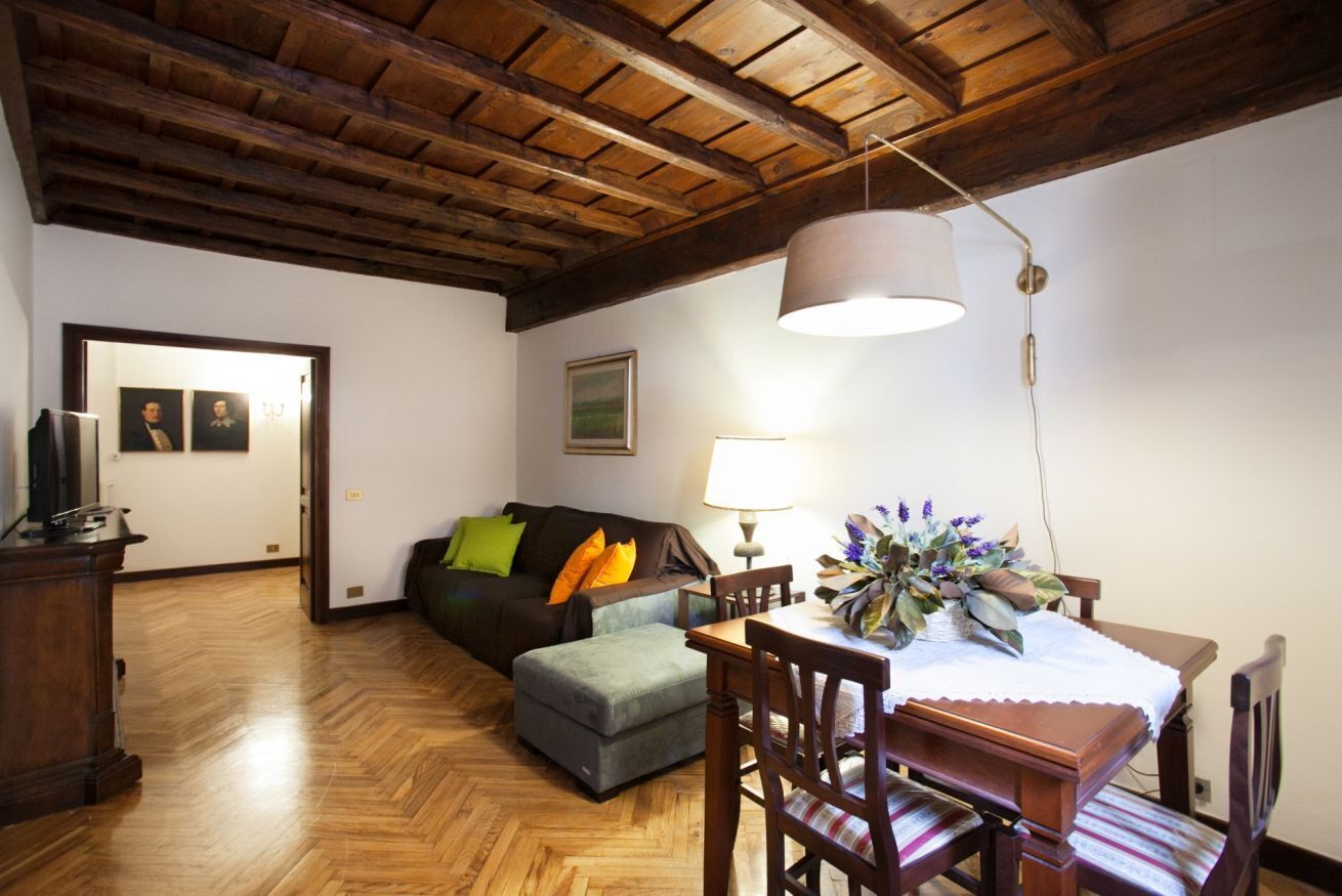 A cozy living room with a wooden ceiling, a sofa, an ottoman, a dining table with chairs, and a lamp.