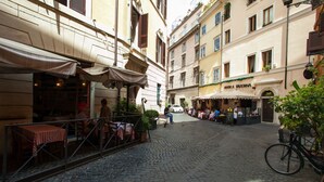 Una strada acciottolata con tavolini all'aperto di un caffè, circondata da edifici con balconi e tende da sole.